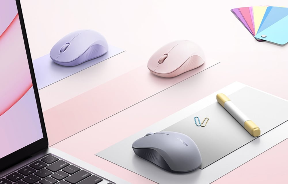 Trzy myszy bezprzewodowe Ugreen M331 w szarym, fioletowym i różowym kolorze stojące na biurku obok laptopa.