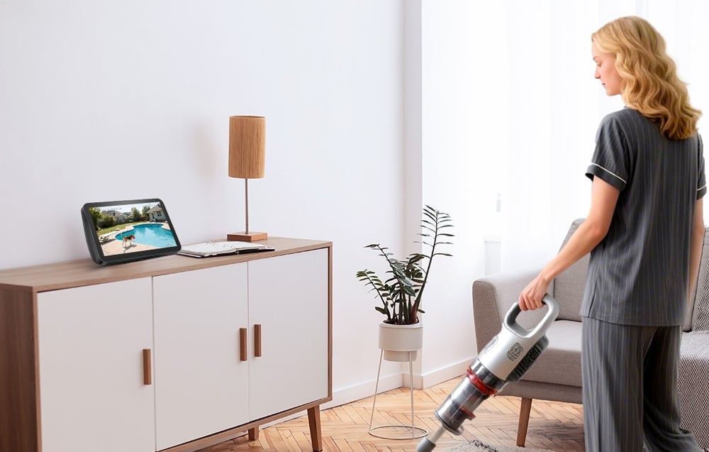Kobieta odkurza salon podczas oglądania obrazu z kamery monitoringu na tablecie ustawionym na komodzie