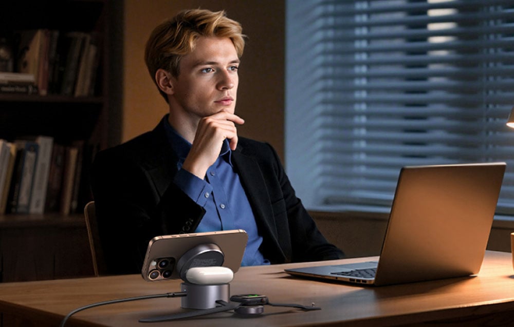 Mężczyzna siedzi przy biurku z laptopem. Obok leżą słuchawki, smartfon i smartwatch podłączone do ładowarki bezprzewodowej.