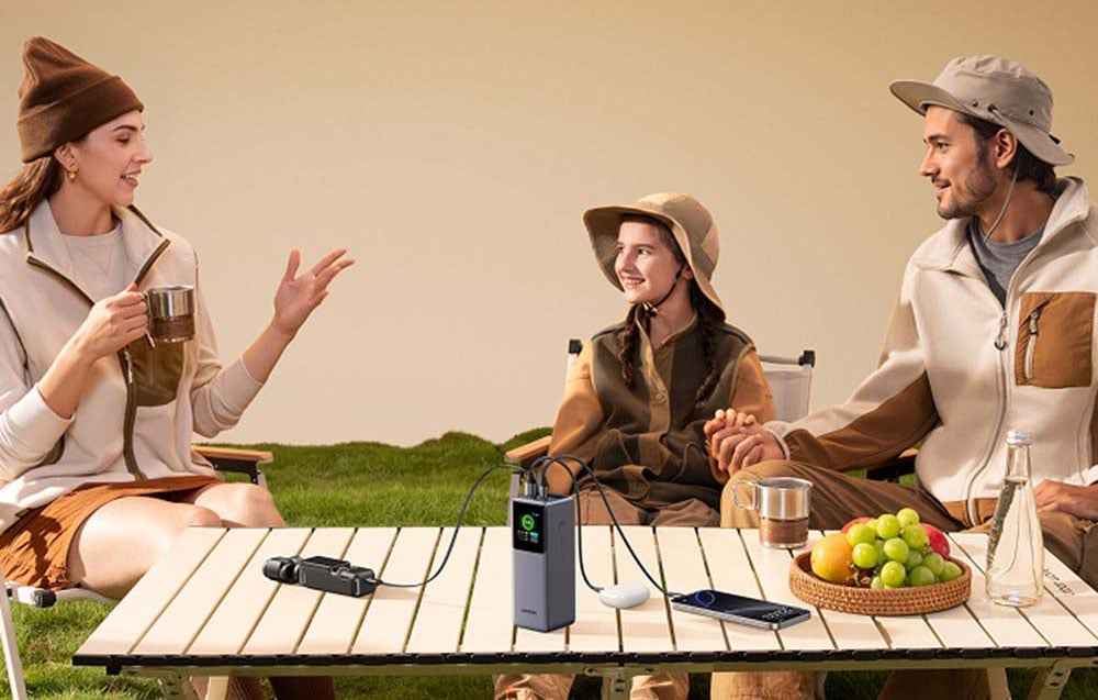 Eine dreiköpfige Familie sitzt beim Picknick an einem Tisch. Auf dem Tisch sind Kopfhörer, ein Smartphone und eine Sportkamera, die an den Powerbank angeschlossen sind.
