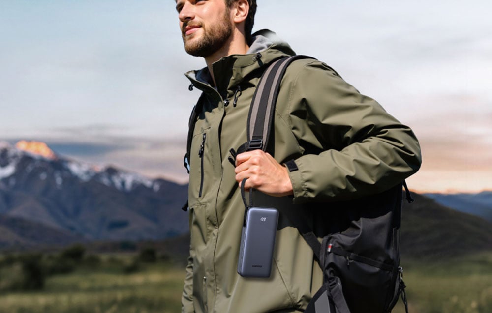 A man in a green jacket with a backpack on his shoulder holding a gray power bank. In the background, a mountain landscape.