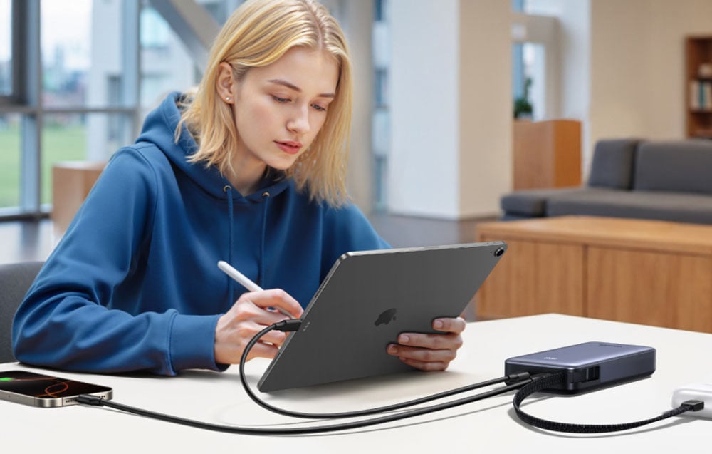 A young woman with blonde hair in a blue sweatshirt sitting at a table holding a tablet and stylus. On the table, there is a power bank connected to a smartphone, wireless headphones, and a tablet.