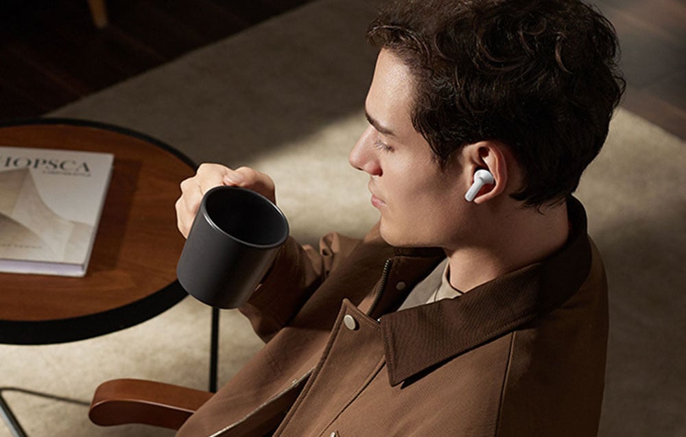 Mężczyzna siedzi w fotelu, trzymając czarny kubek i nosząc białą bezprzewodową słuchawkę douszną