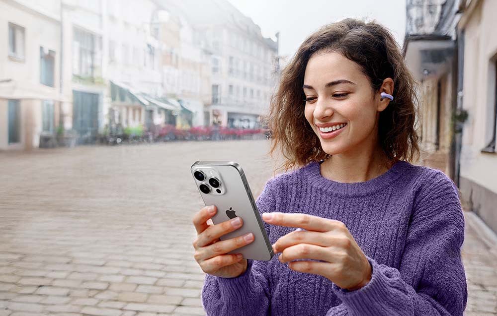 A woman in a purple sweater smiles while looking at her smartphone and wearing a purple wireless earpiece