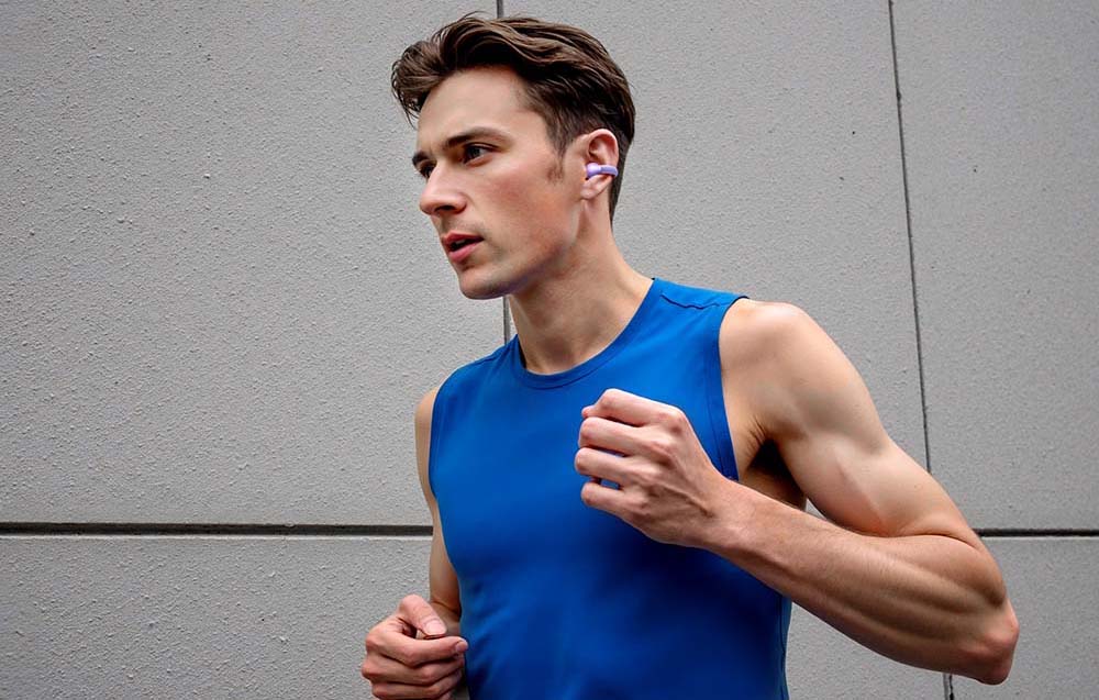 A man in a blue sports top runs by a wall wearing a purple wireless headphone