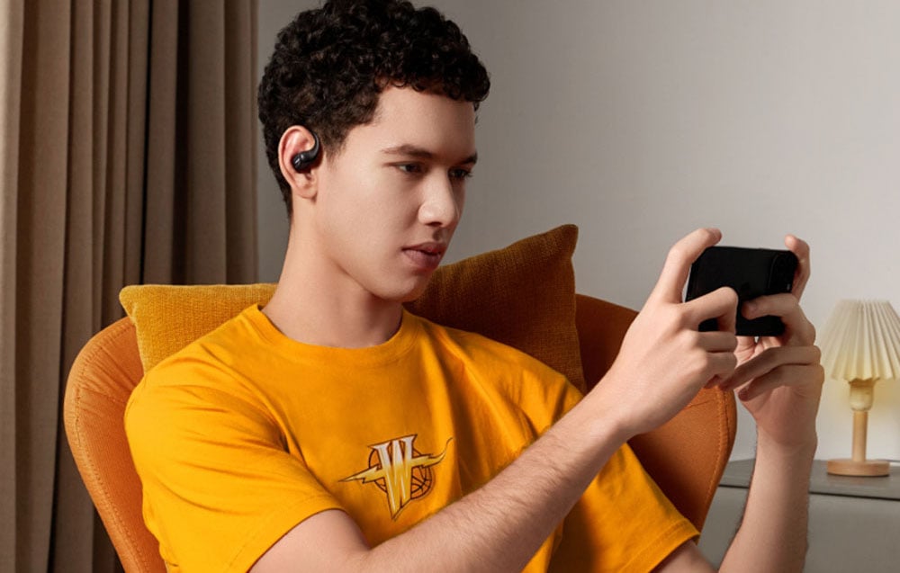 Mężczyzna siedzi w fotelu i gra na smartfonie, mając w uchu czarną słuchawkę bezprzewodową