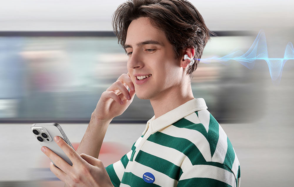 Mężczyzna w zielono-białej koszulce używa telefonu i dotyka słuchawki w uchu, stojąc na tle przejeżdżającego pociągu