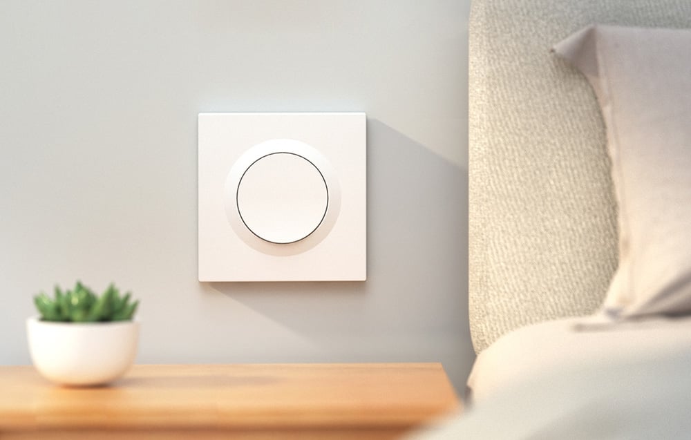 Biały przełącznik zamontowany na ścianie obok łóżka nad szafką nocną z zieloną rośliną.