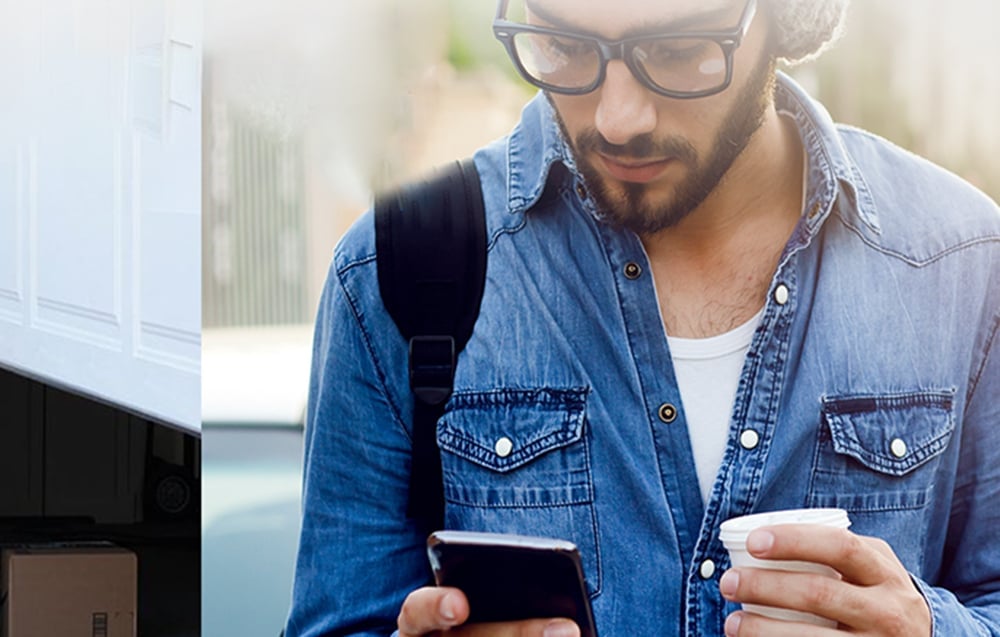 Mężczyzna w okularach patrzący na smartfon, obok fragment otwierającej się bramy garażowej z widoczną paczką