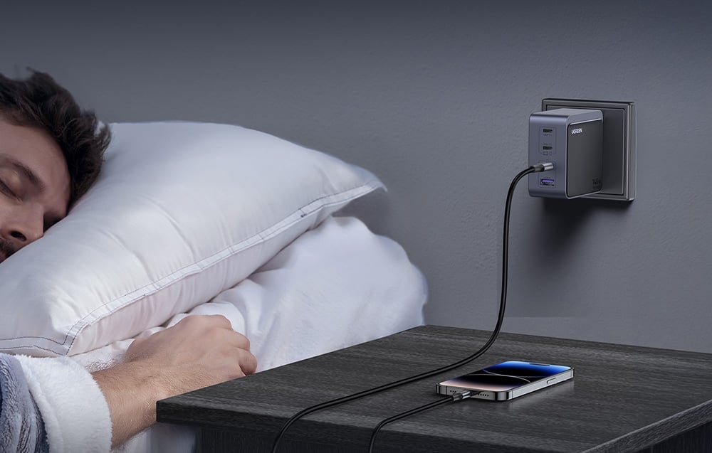 Ładowarka UGREEN Nexode S 140W wpięta do gniazdka ściennego przy łóżku, ładująca smartfon leżący na szafce nocnej obok śpiącego mężczyzny