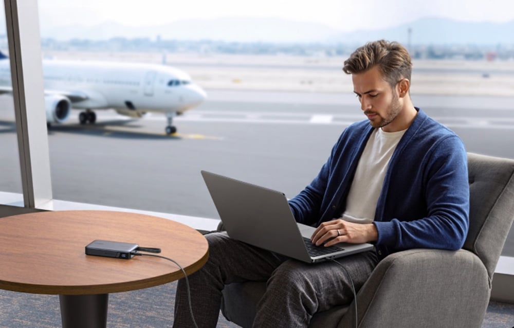 Ein Mann sitzt am Flughafen mit einem Laptop auf den Knien. Der Laptop ist an den Powerbank angeschlossen