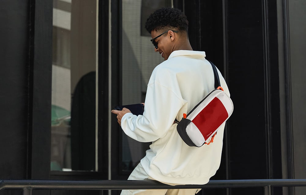 A man in a white hoodie with a red and white waist bag slung over his shoulder near a modern building
