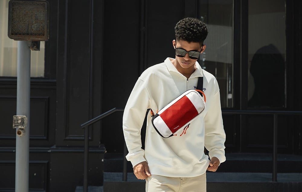 A man in a white hoodie and glasses with a red and white waist bag on his chest in an urban setting
