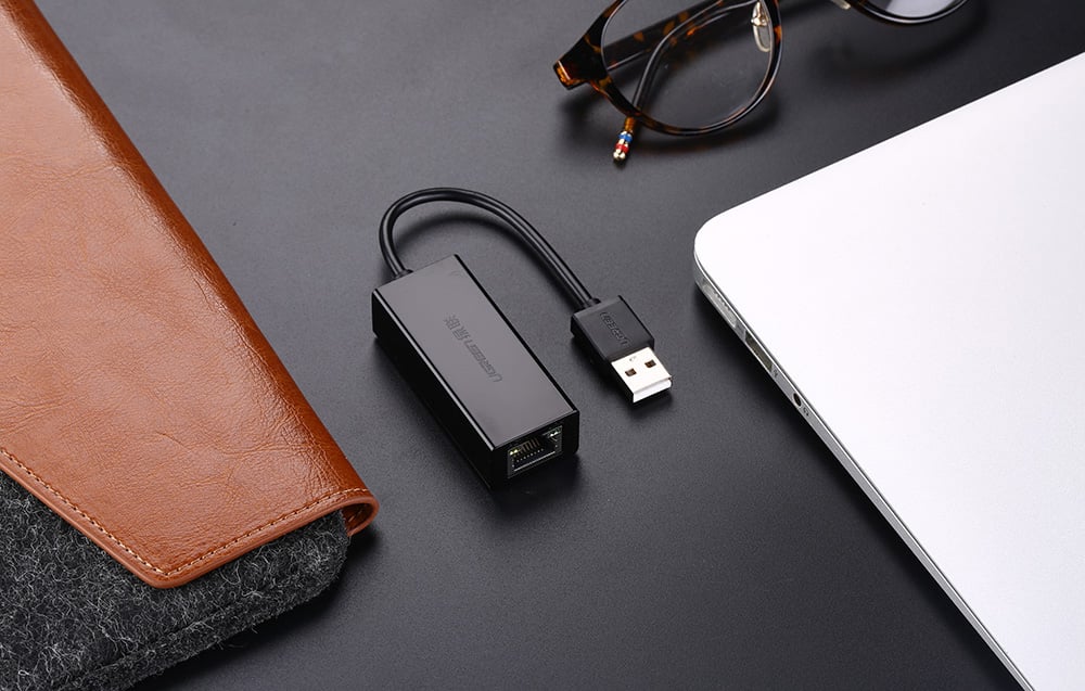 USB adapter with Ethernet port lying on a desk next to a laptop, glasses, and a brown case