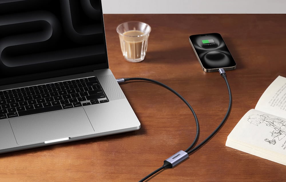 Laptop und Smartphone werden mit einem USB-C Kabel auf einem Holzschreibtisch neben Kaffee und einem Buch geladen