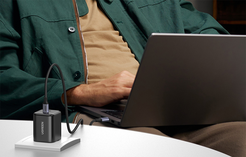 Man in a green jacket working on a laptop with the UGREEN charger connected on a white desk