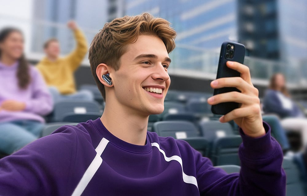 Młody mężczyzna w fioletowej bluzie siedzi na trybunach, uśmiecha się i trzyma smartfon, mając w uchu bezprzewodową słuchawkę