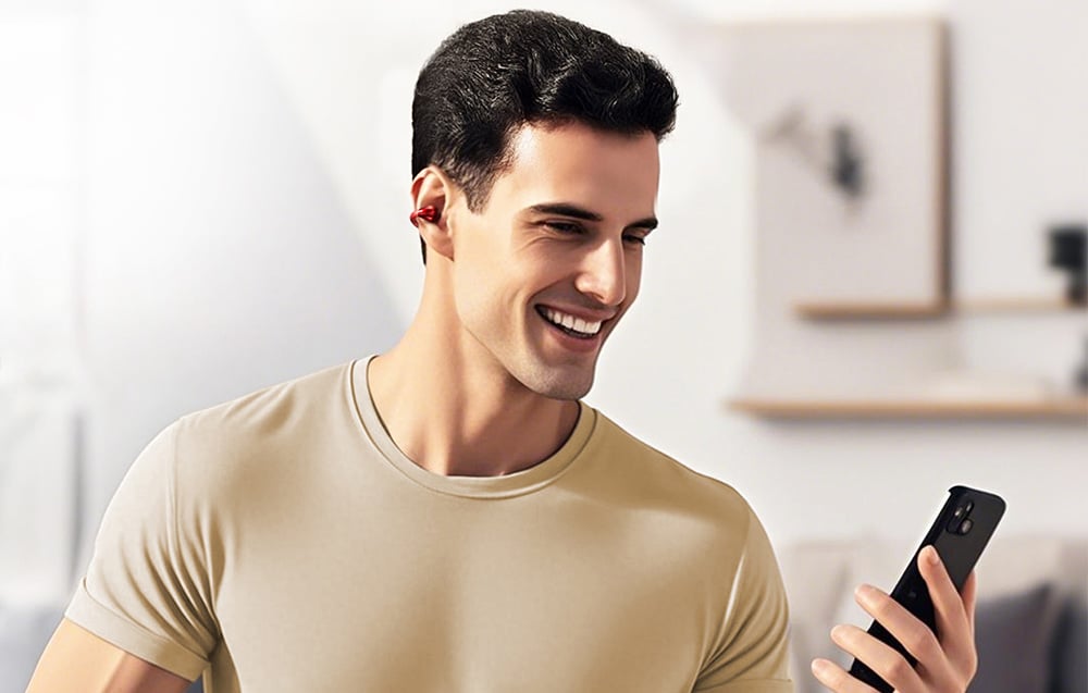 Mężczyzna w beżowej koszulce uśmiecha się, patrząc na telefon, mając czerwoną słuchawkę w uchu