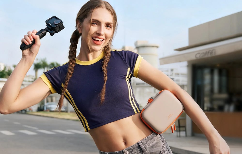 Uśmiechnięta kobieta trzyma kamerę sportową na selfie sticku i beżową torbę Telesin pod pachą