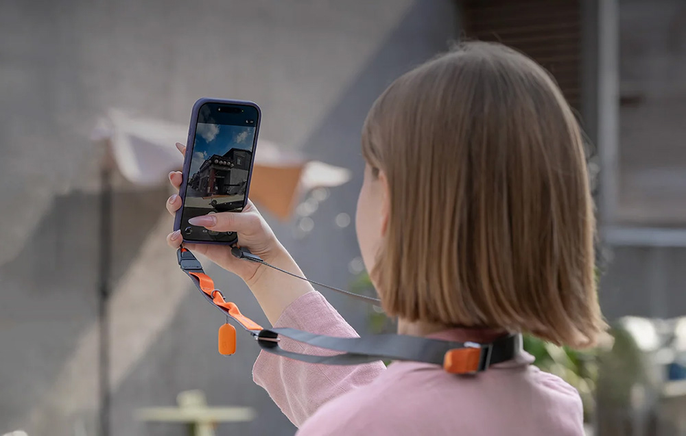 A woman with short hair photographing a building with her phone attached to a neck strap