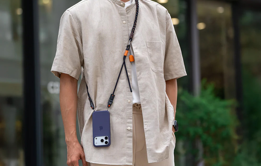 A man in a cream shirt wearing a black strap around his neck with a smartphone attached