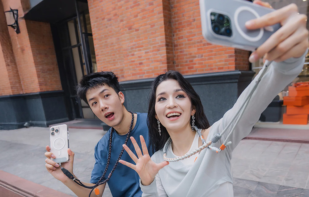 A woman and a man taking a selfie against a brick building, both wearing the PGYTECH strap around their neck