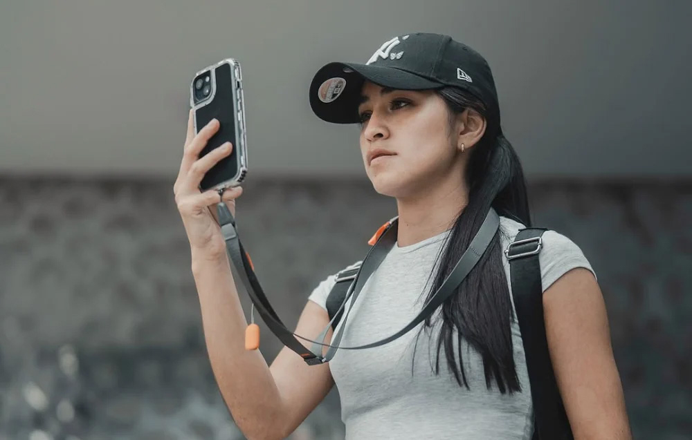 A woman wearing a cap using a phone that is attached to the neck strap