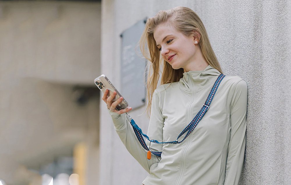 A woman leaning against a wall using her smartphone, which is attached to the neck strap