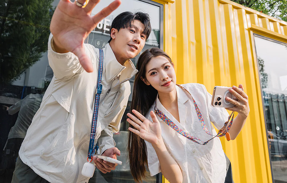 A man and a woman standing next to each other against a yellow building, wearing PGYTECH straps around their necks