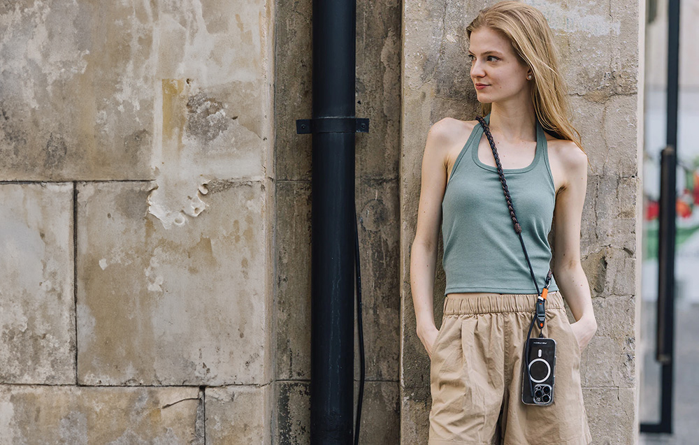 A woman leaning against a stone building, wearing a green tank top and a PGYTECH neck strap