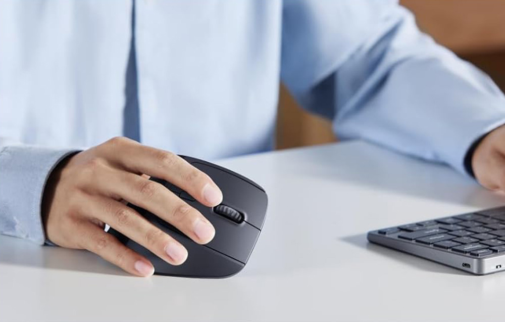 Osoba w niebieskiej koszuli trzyma w dłoni czarną mysz pionową, obok klawiatura na biurku