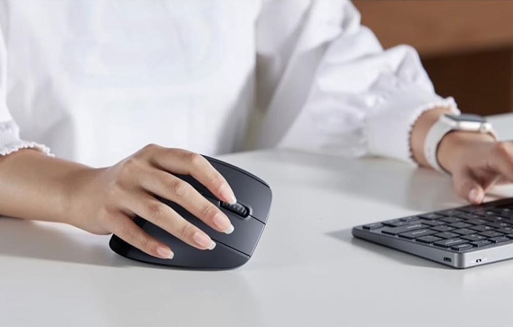 Kobieta w białej bluzce trzyma w dłoni czarną mysz bezprzewodową, drugą dłoń opiera o klawiaturę