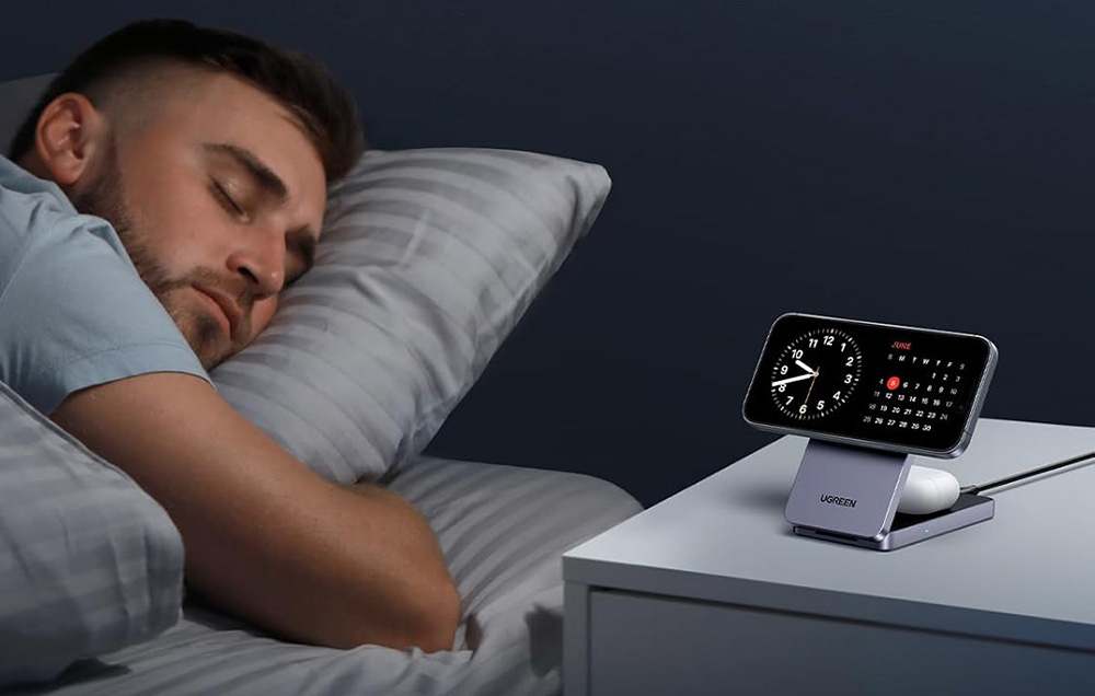 Słuchawki i smartfon podłączone do ładowarki magnetycznej, obok na łóżku śpi mężczyzna