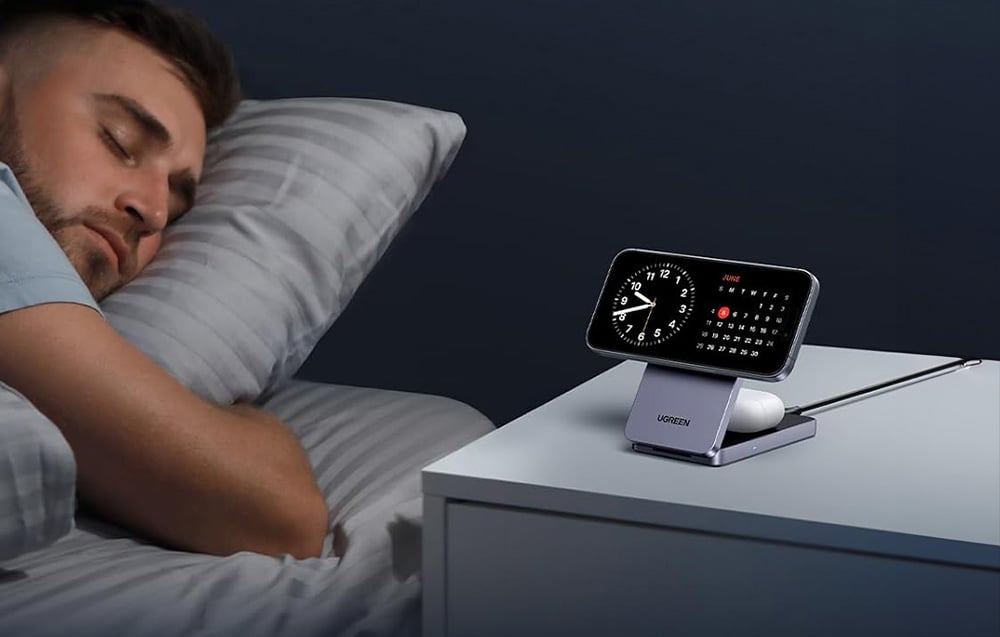 Smartfon i słuchawki TWS podłączone do ładowarki Ugreen na szafce nocnej, obok śpi mężczyzna