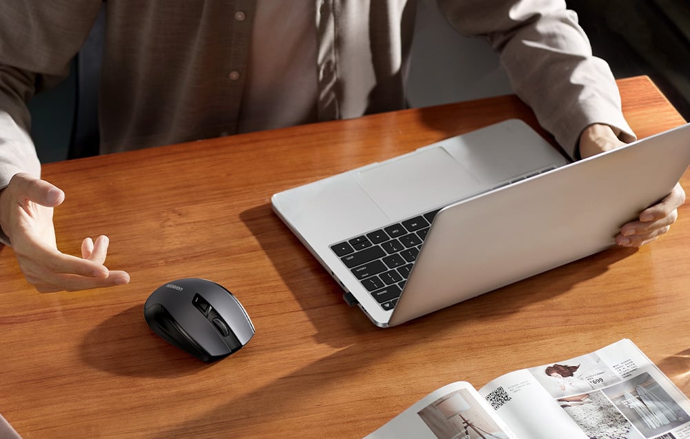 Mężczyzna pracujący przed laptopem, obok na drewnianym biurku czarna mysz Ugreen i katalog z akcesoriami do domu