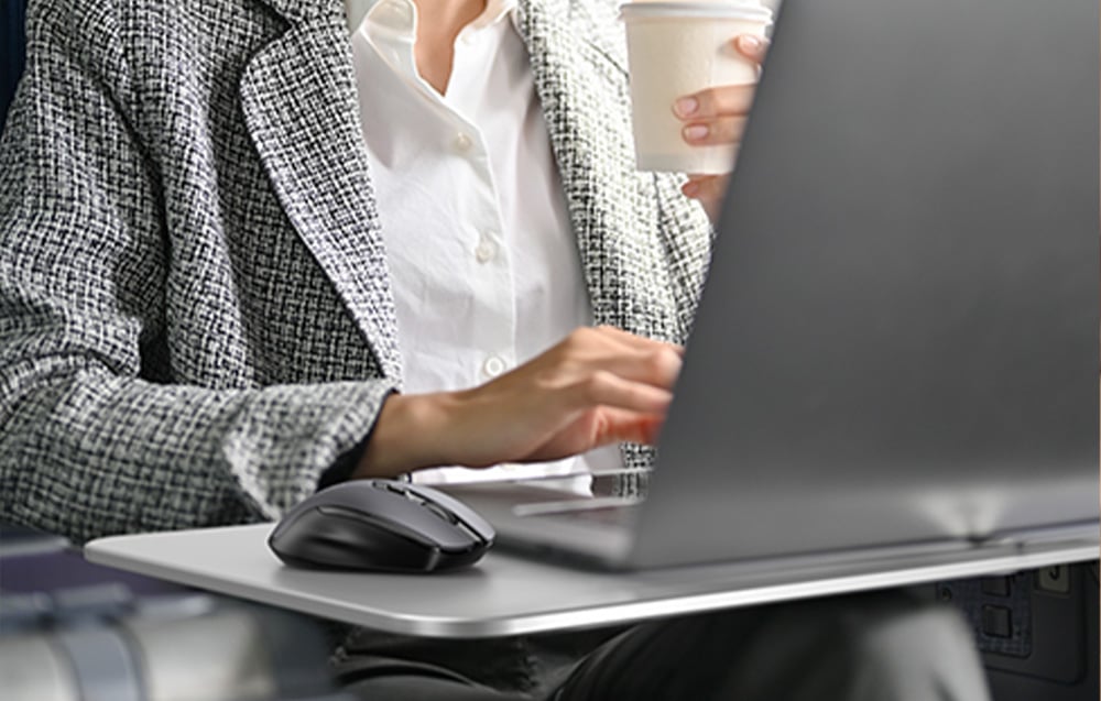 Kobieta w białej koszuli i szarej marynarce pracuje przed laptopem, obok czarna mysz bezprzewodowa