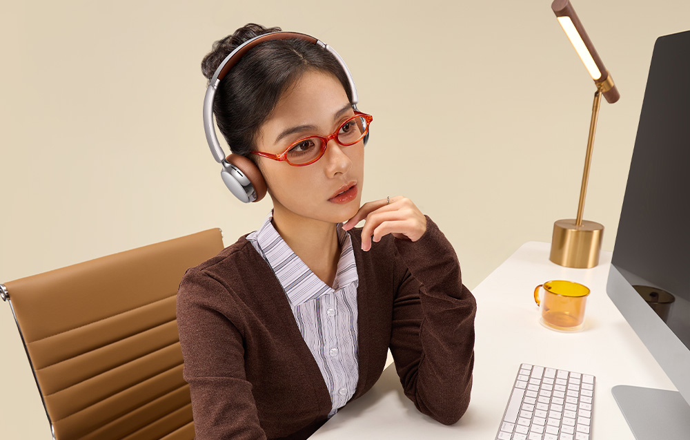 Woman in glasses and over-ear headphones sitting at a desk looking at a monitor
