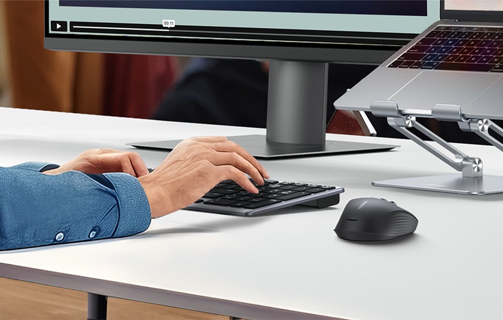 Osoba w niebieskiej koszuli pracuje przed komputerem, obok czarna mysz bezprzewodowa