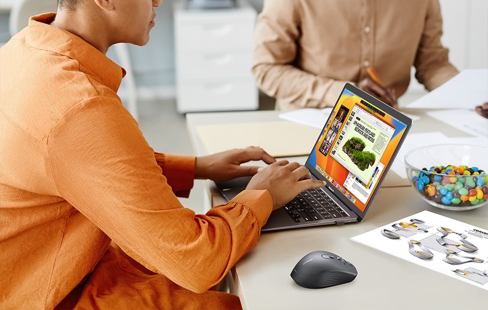 2 osoby siedzące przy stole, jedna z nich używa laptopa i myszy, druga podpisuje dokumenty