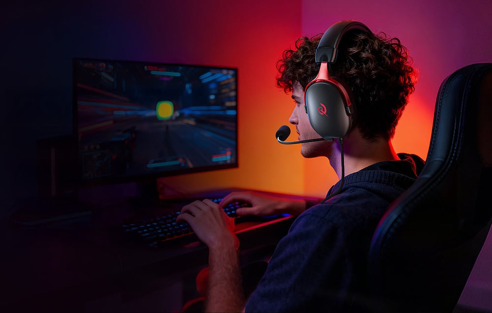 Mężczyzna w słuchawkach z mikrofonem grający na komputerze w ciemnym pokoju oświetlonym kolorowym światłem.