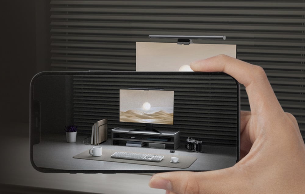 Osoba trzymająca smartfon z widokiem stanowiska komputerowego z monitorem i lampką