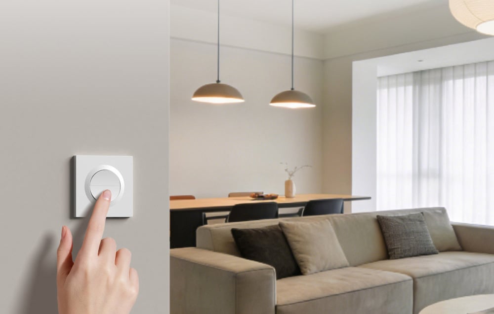 Biały okrągły przełącznik SONOFF zamontowany na jasnej ścianie w nowoczesnym salonie z sofą i jadalnią w tle