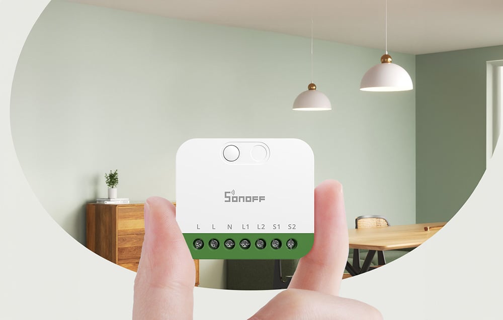 Biały inteligentny przełącznik Sonoff MINI-2GS trzymany w dłoniach, pokazujący terminal z oznaczeniami połączeń L, N, L1, L2, S1, S2, na tle nowoczesnego salonu z lampami wiszącymi