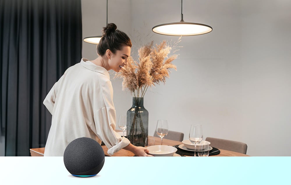 Kobieta układająca nakrycia przy stole z tacą, obok czarnego głośnika inteligentnego Amazon Echo na blacie
