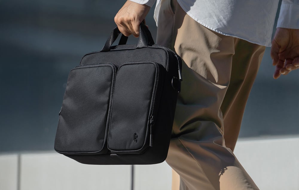 Mężczyzna w koszuli i brązowych spodniach niesie czarną torbę na laptopa