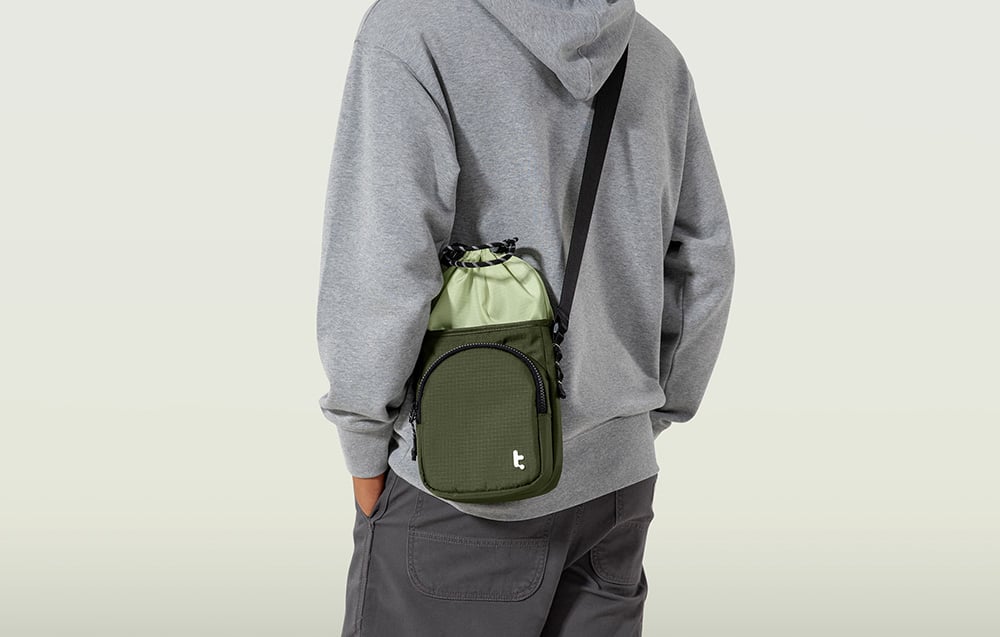 A man in a gray hoodie and dark pants with a green bag slung over his shoulder