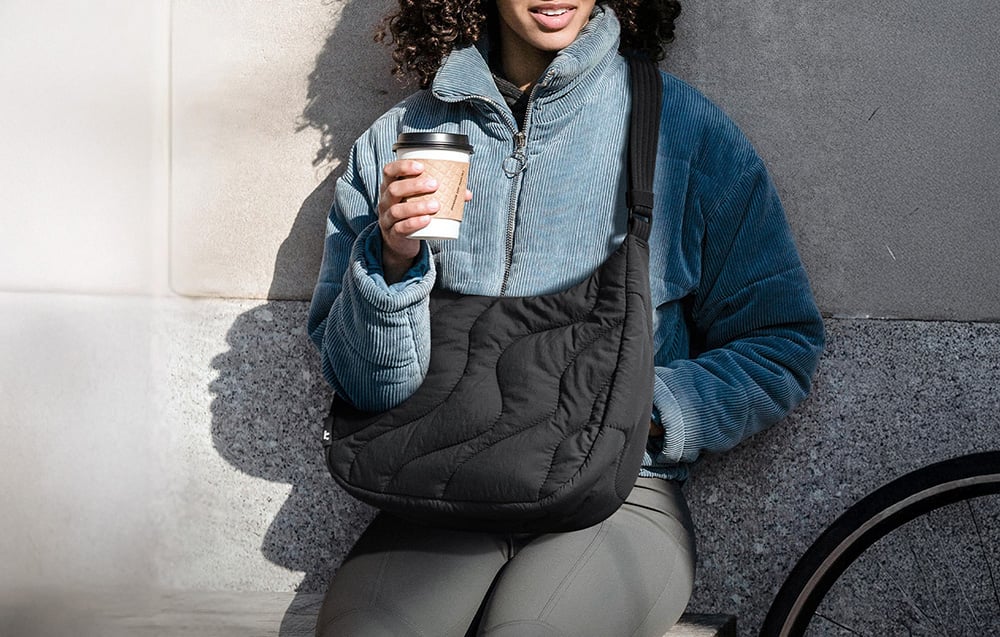 Kobieta z torebką na ramieniu trzyma kubek w dłoni i siedzi na ławce oparta o ścianę budynku