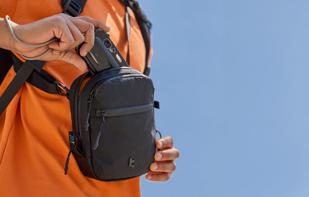 A man in an orange shirt with a bag on his shoulder, taking out a camera