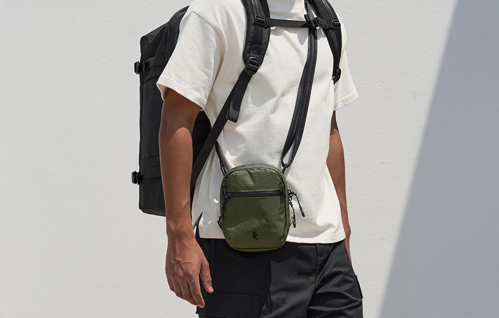 Man in a white shirt with a backpack on his back and a green shoulder bag slung over his shoulder