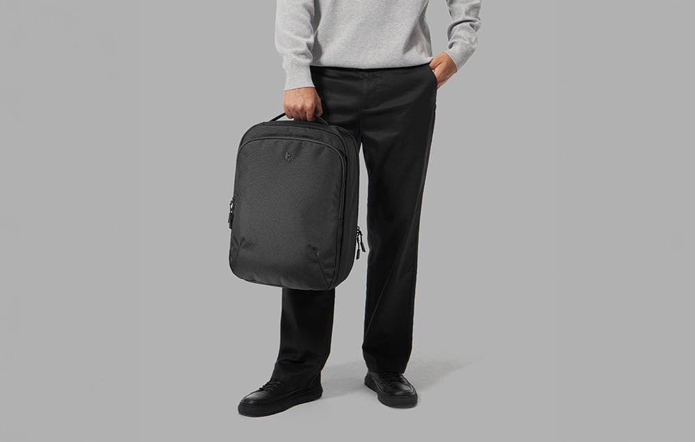 A man in a gray hoodie and black pants holding a laptop backpack in his hand